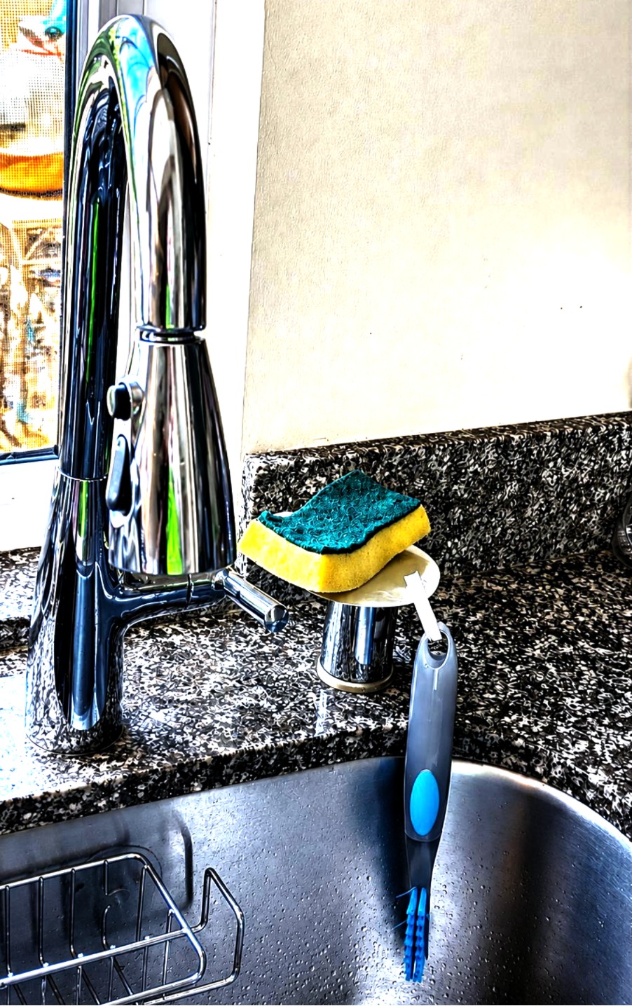 Soap stand mounted over the kitchen air gap beside a chrome faucet, holding a sponge on its tray with a dish brush hanging from the side hook